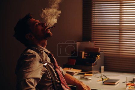 Photo for Young relaxed man with cigarette in mouth blowing smoke while sitting by workplace in office and having break after overtime work - Royalty Free Image