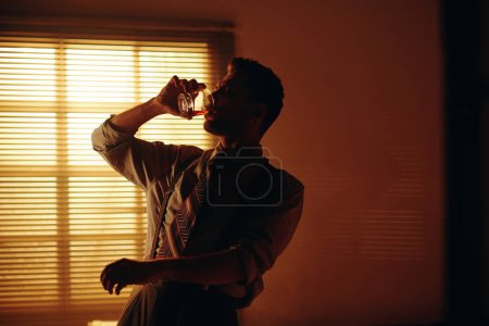 Téléchargez les photos : Jeune homme en formalwear rétro buvant du whisky devant la caméra tout en travaillant tard dans le bureau sombre avec fenêtre couverte de stores vénitiens - en image libre de droit