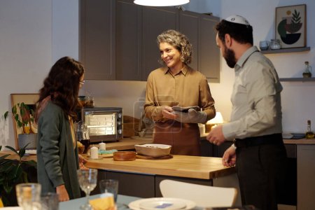 Téléchargez les photos : Heureuse femme juive mature regardant sa fille dans la cuisine tout en étant debout à table avec de la nourriture maison et des bougies - en image libre de droit