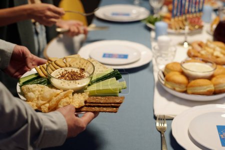 Téléchargez les photos : Mettre l'accent sur l'assiette avec concombre frais, matzo, pain pita et humus dans un petit bol en verre tenu par le jeune homme juif table de service pour le dîner - en image libre de droit