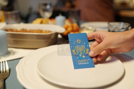 Téléchargez les photos : Main de fille tenant la carte de vœux Hanoukka bleue avec des symboles de fête juive traditionnelle sur une assiette tout en servant la table pour le dîner - en image libre de droit