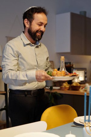 Téléchargez les photos : Un homme barbu souriant tenant une assiette avec une bouteille d'huile et des collations faites maison tout en servant une table pour le dîner Hanoukka dans la cuisine - en image libre de droit