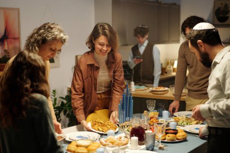 Foto de Familia judía feliz poniendo platos con comida casera en la mesa mientras se sirve para invitados antes de la cena de Hanukkah - Imagen libre de derechos