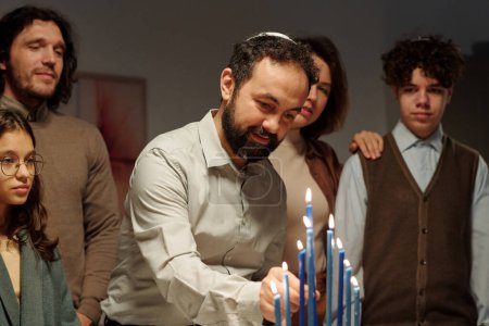 Téléchargez les photos : Homme mûr dans skullcap juif mettant bougie bleue brûlante sur chandelier menorah tout en célébrant Hanoukka à la maison avec sa famille - en image libre de droit