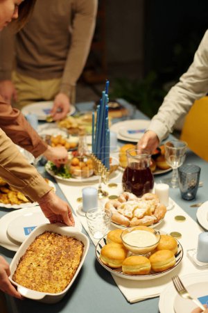 Foto de Manos de miembros de la familia judía poniendo cubiertos con comida casera apetitosa en la mesa servida para la cena de Hanukkah - Imagen libre de derechos