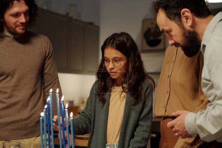 Photo for Cute Jewish girl lighting candles on menorah candlestick for Hanukkah while standing among young and mature men during celebration - Royalty Free Image