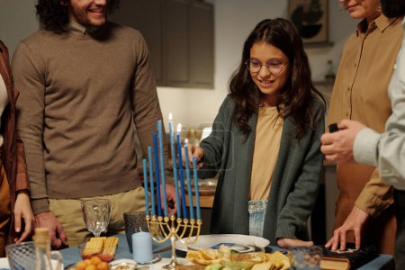 Foto de Linda chica sonriente de la familia judía encendiendo velas menorah mientras está de pie entre su hermano mayor y su madre - Imagen libre de derechos