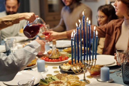 Photo for Focus on blue burning candles on menorah candlestick standing on table served with homemade food and drinks during Hanukkah family dinner - Royalty Free Image