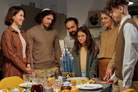 Foto de Familia judía feliz de tres generaciones mirando velas de menorah encendidas de pie sobre la mesa servida entre comida y bebidas caseras - Imagen libre de derechos
