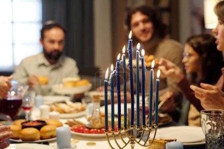 Photo for Burning candles on menorah candlestick standing on served table with homemade food and drinks against members of Jewish family - Royalty Free Image