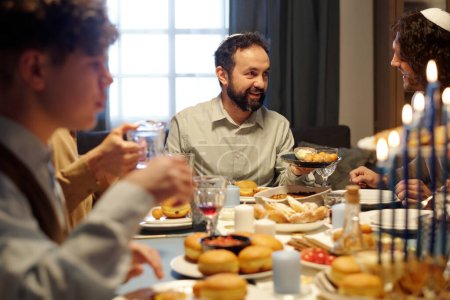 Téléchargez les photos : Focus sur l'homme juif barbu mature offrant à son fils ou invité essayer pâtisserie traditionnelle maison tout en tenant assiette avec des beignets savoureux - en image libre de droit
