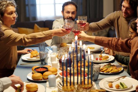 Foto de Miembros de una gran familia judía tintinean con gafas de vino sobre la mesa servida con comida casera mientras brindan por Hanukkah para la cena. - Imagen libre de derechos