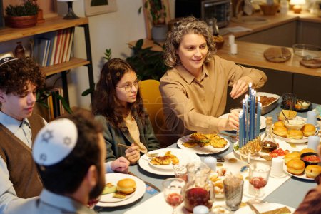 Téléchargez les photos : Femme souriante allumant des bougies menorah sur le chandelier debout sur la table servie parmi la nourriture et les boissons faites maison pendant le dîner - en image libre de droit