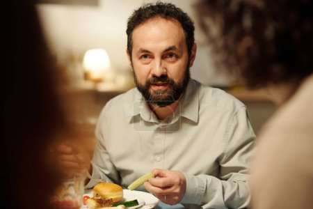 Téléchargez les photos : Homme juif mature avec un morceau de concombre frais à la main regardant invité ou membre de la famille pendant le chat par Hanoukka dîner - en image libre de droit
