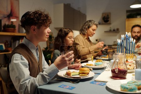Téléchargez les photos : Vue latérale du garçon juif aux cheveux bouclés tenant un verre de boisson maison et ayant un beignet et d'autres aliments Hanoukka pendant le dîner festif - en image libre de droit