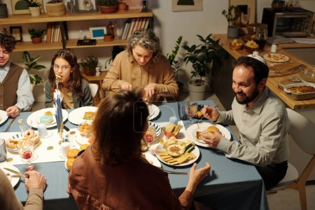 Téléchargez les photos : Angle élevé de la famille juive réunie autour d'une table servie ayant de la nourriture maison et discutant de quelque chose pendant la célébration de Hanoukka - en image libre de droit