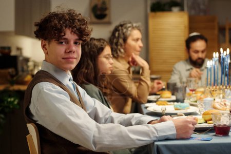 Téléchargez les photos : Jeune garçon juif en tenue décontractée regardant la caméra à table servie avec de la nourriture et des boissons maison contre des membres de sa famille - en image libre de droit