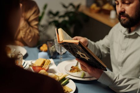 Foto de Primer plano del hombre judío maduro sosteniendo abierto el Antiguo Testamento sobre la mesa servida y leyendo Salmos a los miembros de su familia durante la cena - Imagen libre de derechos
