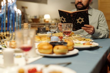 Photo for Focus on cropped mature man reading Torah in black cover with star of David while sitting by festive table served with homemade food - Royalty Free Image