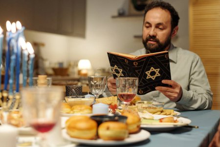 Photo for Mature man with open Torah with star of David on cover reading Psalms or some other texts from Old Testament during Hanukkah dinner - Royalty Free Image