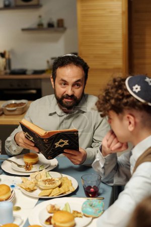 Téléchargez les photos : Focus sur l'homme mûr barbu en kippa en regardant son fils pré-adolescent tout en lui expliquant texte de la Torah par dîner Hanoukka festive - en image libre de droit
