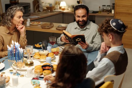Photo for Happy bearded man with open Scripture sitting in front of his family and looking at daughter during discussion of text from Old Testament - Royalty Free Image