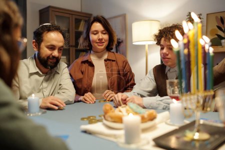 Foto de Varios miembros de la familia judía apostando durante el juego de ocio mientras están sentados a la mesa con velas encendidas en el candelero menorah - Imagen libre de derechos