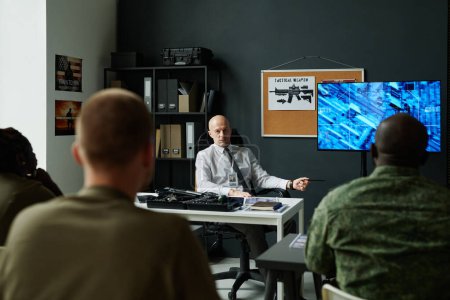 Photo for Confident mature officer sitting by workplace and making presentation of military tactics on interactive screen to group of students - Royalty Free Image