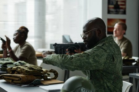 Téléchargez les photos : Afro-Américain en uniforme de camouflage tirer à partir d'une arme pendant l'entraînement militaire tout en étant assis contre ses camarades de classe - en image libre de droit