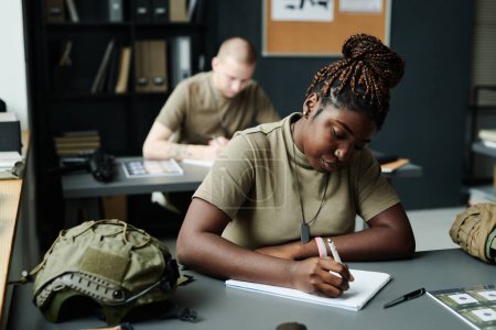 Foto de Joven estudiante afroamericana tomando notas en bloc de notas después del profesor mientras está sentada junto al escritorio en la lección de entrenamiento militar - Imagen libre de derechos