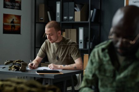 Photo for Young man in t-shirt sitting by desk and making notes in copybook after teacher while working at lesson of military training - Royalty Free Image
