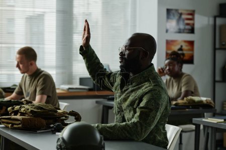 Foto de Chico afroamericano en uniforme militar y anteojos levantando la mano y mirando al profesor después de una conferencia o presentación para hacer una pregunta - Imagen libre de derechos