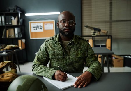 Photo for Young serious African American man in camouflage uniform sitting by desk, making notes and listening to explanation of teacher - Royalty Free Image
