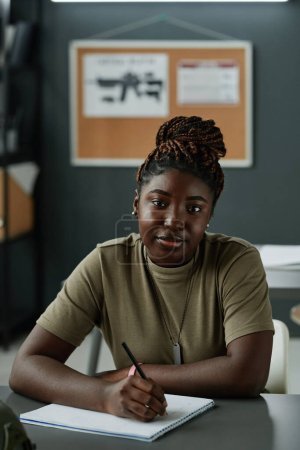 Foto de Joven mujer afroamericana seria con la pluma en la mano mirando a la cámara mientras se sienta junto al escritorio y toma notas de la conferencia - Imagen libre de derechos