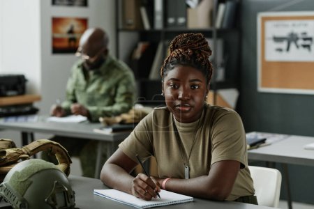 Photo for African American female student in casualwear sitting by desk with military equipment and looking at camera while making lecture notes - Royalty Free Image
