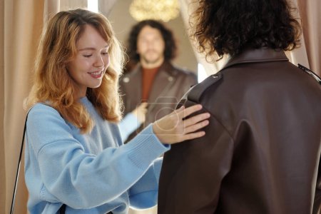 Foto de Joven mujer sonriente de pie junto a su marido probándose una nueva chaqueta de cuero frente al espejo en el probador durante las compras - Imagen libre de derechos