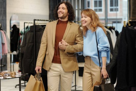 Photo for Young cheerful couple in quiet luxury attire looking at new seasonal collection of clothes and laughing while shopping in boutique - Royalty Free Image
