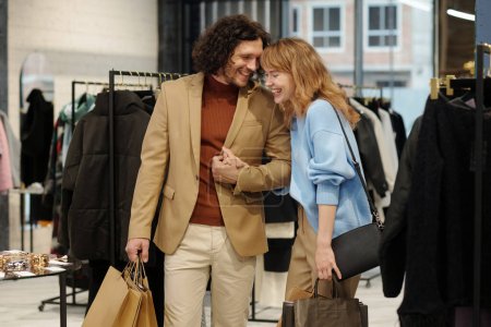 Foto de Alegre joven hombre y mujer riendo mientras está de pie en la boutique entre bastidores con la nueva colección de moda de temporada y pequeña pantalla - Imagen libre de derechos