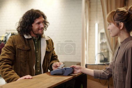 Foto de Joven cliente masculino sonriente mirando al asistente de la tienda bonita mientras está parado junto al mostrador y pagando por la compra con tarjeta de crédito - Imagen libre de derechos