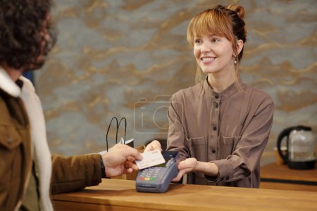 Foto de Asistente de tienda joven feliz mirando al cliente con tarjeta de crédito mientras paga por ropa u otros bienes comprados en la boutique - Imagen libre de derechos