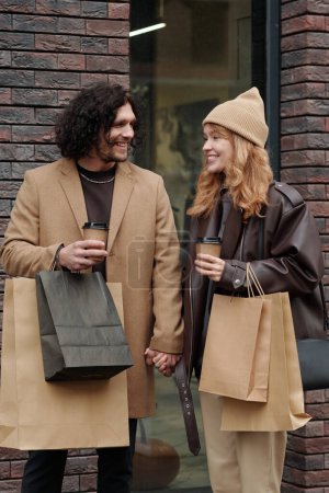 Foto de Joven hombre y mujer sonrientes con café para llevar y bolsas de papel mirándose mientras están de pie junto al edificio de ladrillo - Imagen libre de derechos