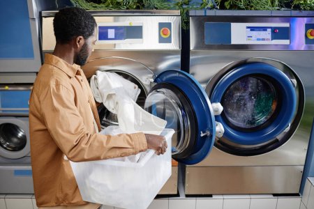 Foto de Vista lateral del joven hombre negro en camisa beige poniendo ropa blanca en el tambor de la lavadora automática mientras está de pie en la lavandería - Imagen libre de derechos