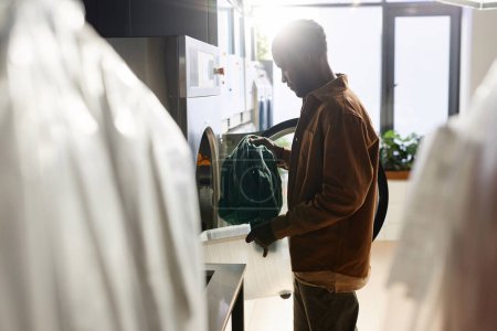 Foto de Vista lateral de los jóvenes afroamericanos cliente masculino de lavandería café de pie delante de la lavadora automática con puerta redonda abierta - Imagen libre de derechos