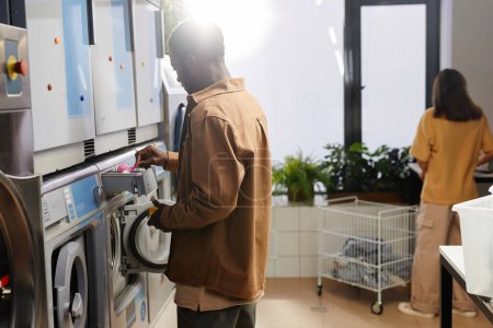 Téléchargez les photos : Jeune homme afro-américain mettant de la poudre à laver dans un petit compartiment spécial dans une machine automatique dans un café à linge - en image libre de droit