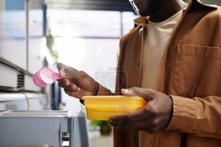Photo for Close-up of young African American man with cap of washing powder pouring it into special compartment in automatic machine - Royalty Free Image