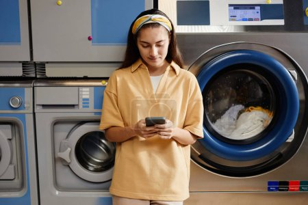 Foto de Mujer bonita en ropa de casualwear mirando la pantalla del teléfono inteligente y mensajes de texto, mientras que de pie contra la lavadora en la cafetería de lavandería - Imagen libre de derechos