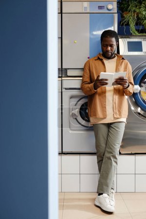 Téléchargez les photos : Jeune homme afro-américain en tenue décontractée debout à côté de la machine à laver et le livre de lecture en couverture blanche dans le café de blanchisserie moderne - en image libre de droit