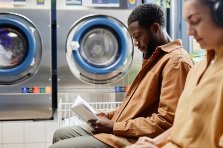 Téléchargez les photos : Sérieux homme afro-américain lecture livre à côté de la jeune femme dans les écouteurs tout en étant assis contre les machines à laver - en image libre de droit