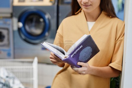 Téléchargez les photos : Plan recadré de livre de lecture fille en couverture violette tout en se tenant devant la caméra contre les machines à laver dans le café de blanchisserie - en image libre de droit