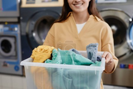 Foto de Foto recortada de mujer joven sosteniendo contenedor de plástico con un montón de ropa limpia en frente de la cámara mientras está de pie en el café de lavandería - Imagen libre de derechos